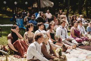 Guests seated outdoors on rugs at a bohemian wedding ceremony, wearing rustic attire in a sunlit garden setting.
