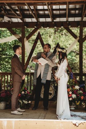 Couple exchanges vows under wooden gazebo with officiant, floral arch backdrop, bride in white gown, groom in brown suit.