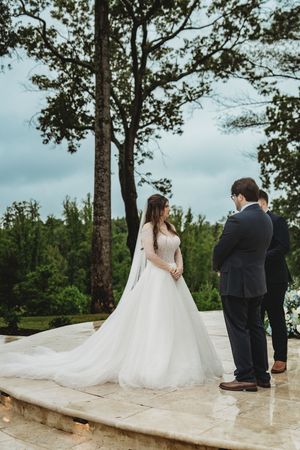 Bride in white ball gown and veil stands with groom in dark suit during outdoor wedding ceremony surrounded by lush trees.