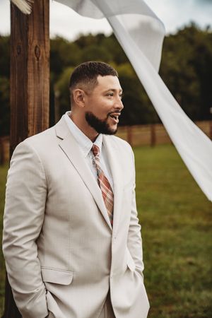 Smiling groom in a beige suit and striped tie stands outdoors at a rustic wedding venue with white fabric decor.
