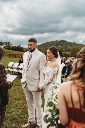 Bride and groom holding hands at outdoor wedding ceremony on green field with guests seated and rolling hills in background.