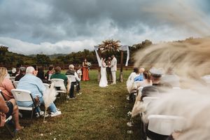 Outdoor wedding ceremony on green lawn with couple at floral arch, guests seated in white chairs under dramatic cloudy sky.