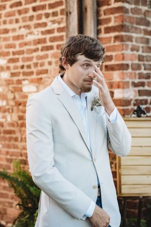 Emotional groom in white blazer with boutonniere wiping tear at outdoor wedding ceremony near brick wall.
