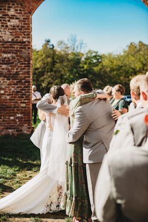 Bride and groom share first kiss at outdoor wedding ceremony near rustic brick arch surrounded by wedding party.