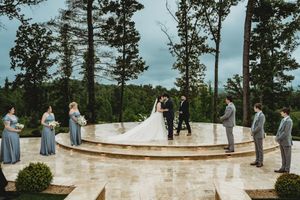 Bride and groom kiss at outdoor wedding ceremony on stone terrace surrounded by bridesmaids and groomsmen with forest backdrop.