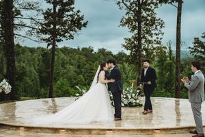 Bride and groom exchange vows at an outdoor wedding ceremony on a stone platform surrounded by tall trees.