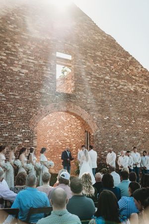 Outdoor wedding ceremony at historic brick ruins with bridal party, officiant, and seated guests in warm sunlight.