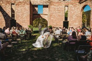 Outdoor wedding ceremony in rustic brick ruins with bride and groom kissing in the aisle surrounded by seated guests.