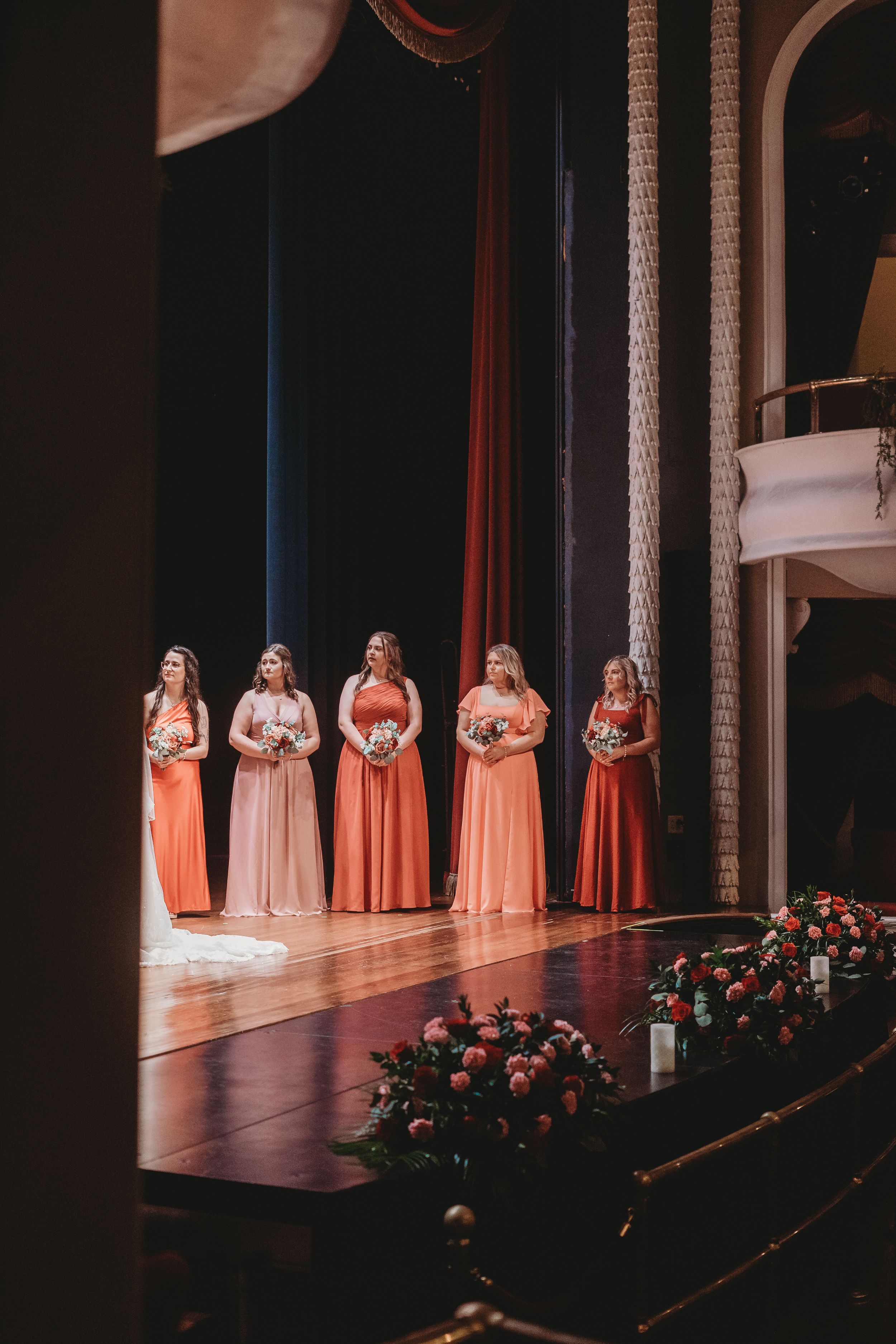 Bridesmaids in coral and blush gowns stand on illuminated stage during wedding ceremony.