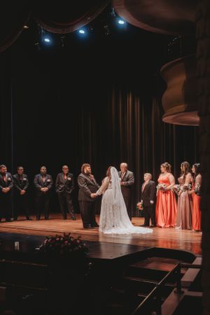 Wedding ceremony on dimly lit stage with bridesmaids in coral dresses and audience in view.