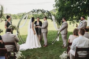 Outdoor wedding ceremony under trees with groomsmen in beige suits and bridesmaids in teal dresses.