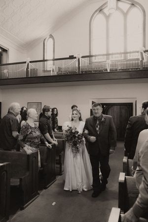 Black and white photo of a bride walking down the church aisle with her escort, guests standing in pews.