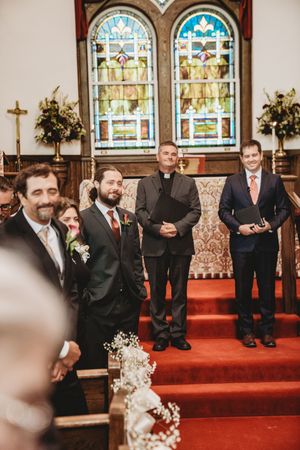 Men in suits stand at a church altar with stained glass windows during a wedding ceremony.