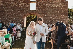 Bride and groom walk down outdoor aisle at rustic brick venue as guests celebrate with confetti.