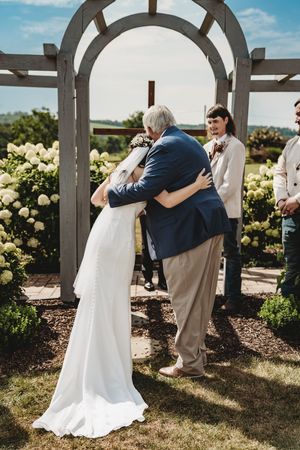 Bride and father share emotional hug at outdoor wedding ceremony under wooden arch with white hydrangeas.