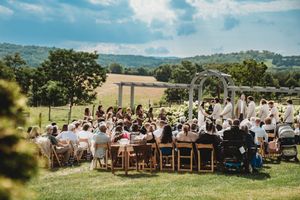 Outdoor wedding ceremony on a green hillside with guests seated in wooden chairs facing a floral arbor under a blue sky.