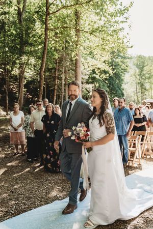 Bride and groom walk down outdoor woodland wedding aisle, bride holding colorful bouquet, guests standing among tall trees.
