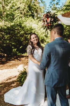 Smiling bride in lace wedding dress holds hands with groom in blue suit during outdoor woodland ceremony.