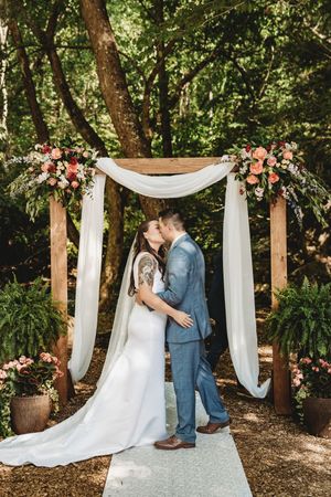 Bride and groom share first kiss under floral wooden arch with white draping in lush forest wedding ceremony.