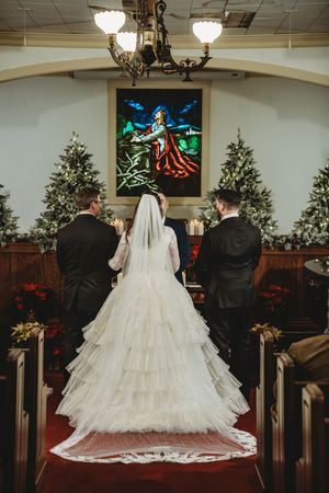 Bride in tiered white gown with veil at church altar flanked by Christmas trees during winter wedding ceremony.