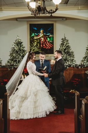 Bride and groom exchange vows at Christmas-decorated church altar with stained glass window and officiant holding Bible.