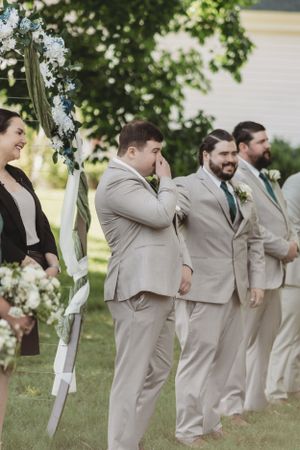 Groomsmen lined up outdoors wearing matching beige suits and ties at a summer wedding.