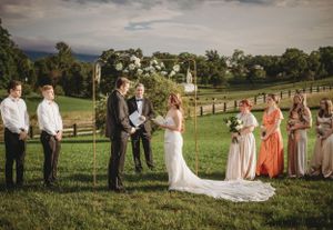Wedding party poses outdoors in a scenic field with rolling hills and wooden fences at sunset.