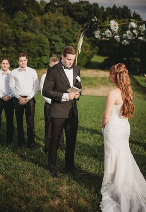 Wedding ceremony taking place in a green field with rustic wooden fence and natural landscape.