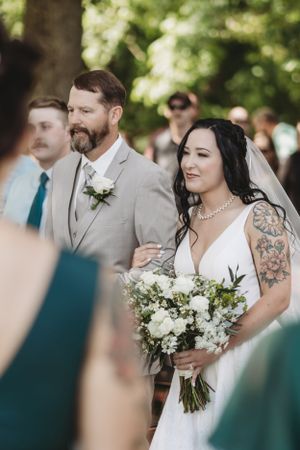 Wedding party members gather outdoors with white floral bouquets and beige suits.