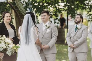 Wedding ceremony participants in beige suits and white dresses gather under trees.