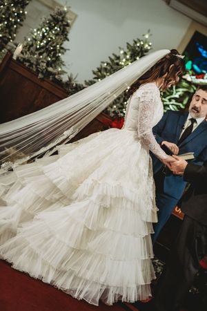 Bride in layered tulle wedding gown with lace sleeves and long veil stands at altar during church ceremony.