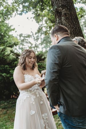 Smiling bride in floral gown exchanges rings with groom outdoors during intimate woodland wedding ceremony.