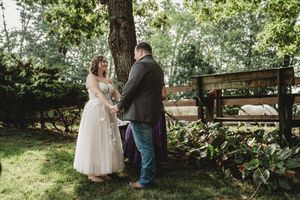 Couple exchanging vows outdoors under a tree, bride in white gown, groom in gray suit, lush green garden wedding ceremony.