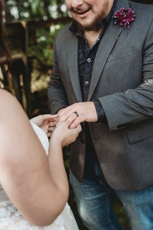 Groom in gray suit placing wedding ring on bride's finger during outdoor ceremony.