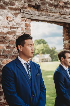 Two groomsmen in navy blue suits and floral ties stand near a rustic brick wall at an outdoor wedding ceremony.