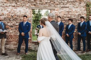 Bride in white gown and veil walks with older man past groomsmen in navy suits at outdoor brick wall ceremony.