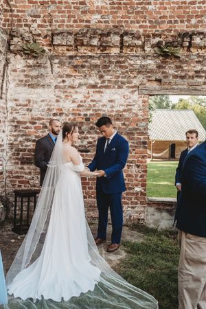 Bride and groom exchange vows at rustic brick wall outdoor wedding ceremony with officiant and groomsman present.