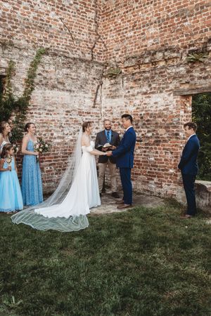 Bride and groom exchange vows outdoors before a rustic brick wall, with bridesmaids in blue dresses and an officiant present.