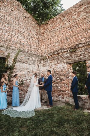 Outdoor wedding ceremony in historic brick ruins with bride in white gown, groom in navy suit, officiant, and wedding party.
