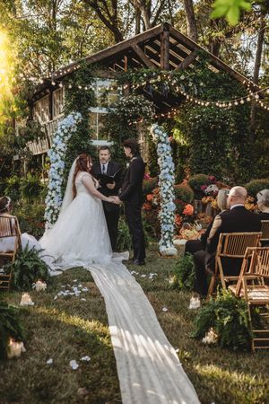 Outdoor garden wedding ceremony with bride and groom at floral arch, string lights, and guests seated on wooden chairs.