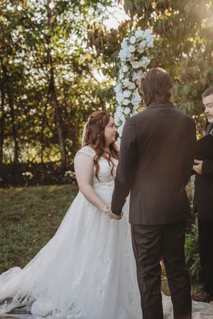 Bride in white lace gown holds hands with groom in dark suit during outdoor wedding ceremony with floral arch.