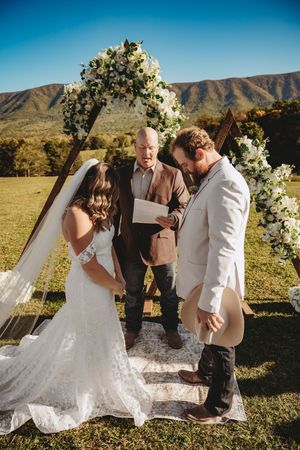 Outdoor wedding ceremony with bride and groom at floral arch, officiant reading vows, mountain backdrop on sunny day.