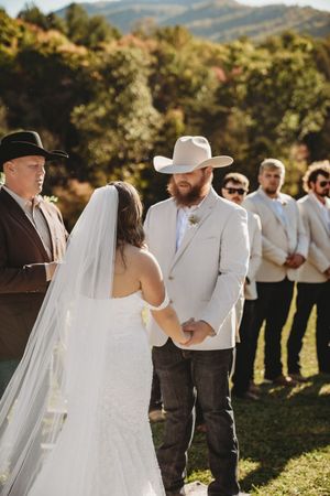 Outdoor wedding ceremony with groom in white cowboy hat and suit holding hands with bride in white veil, mountains in background.