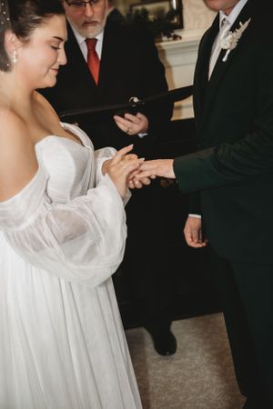 Wedding couple exchanges rings during indoor ceremony surrounded by formal attire.