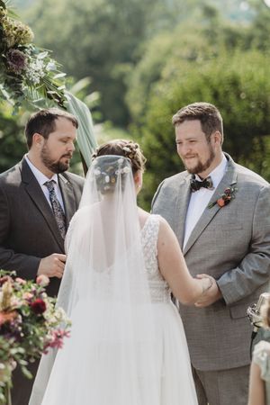 Bride and groom exchange vows outdoors, officiant beside them, lush greenery backdrop, floral arch, wedding ceremony.