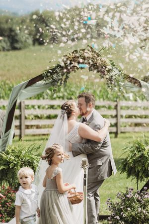 Bride and groom share a kiss under floral arch with confetti, flower girl and ring bearer nearby at outdoor wedding.