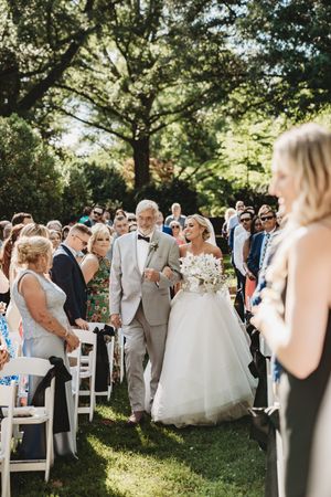 Bride walks down outdoor aisle with older gentleman in gray suit, surrounded by seated guests under lush green trees.