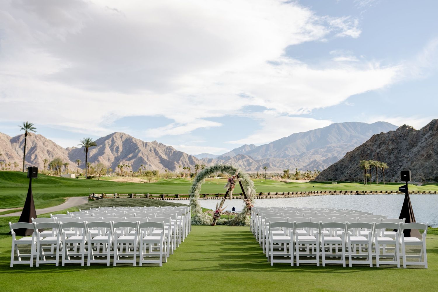 Outdoor wedding ceremony setup with white chairs and floral arch against mountain backdrop.