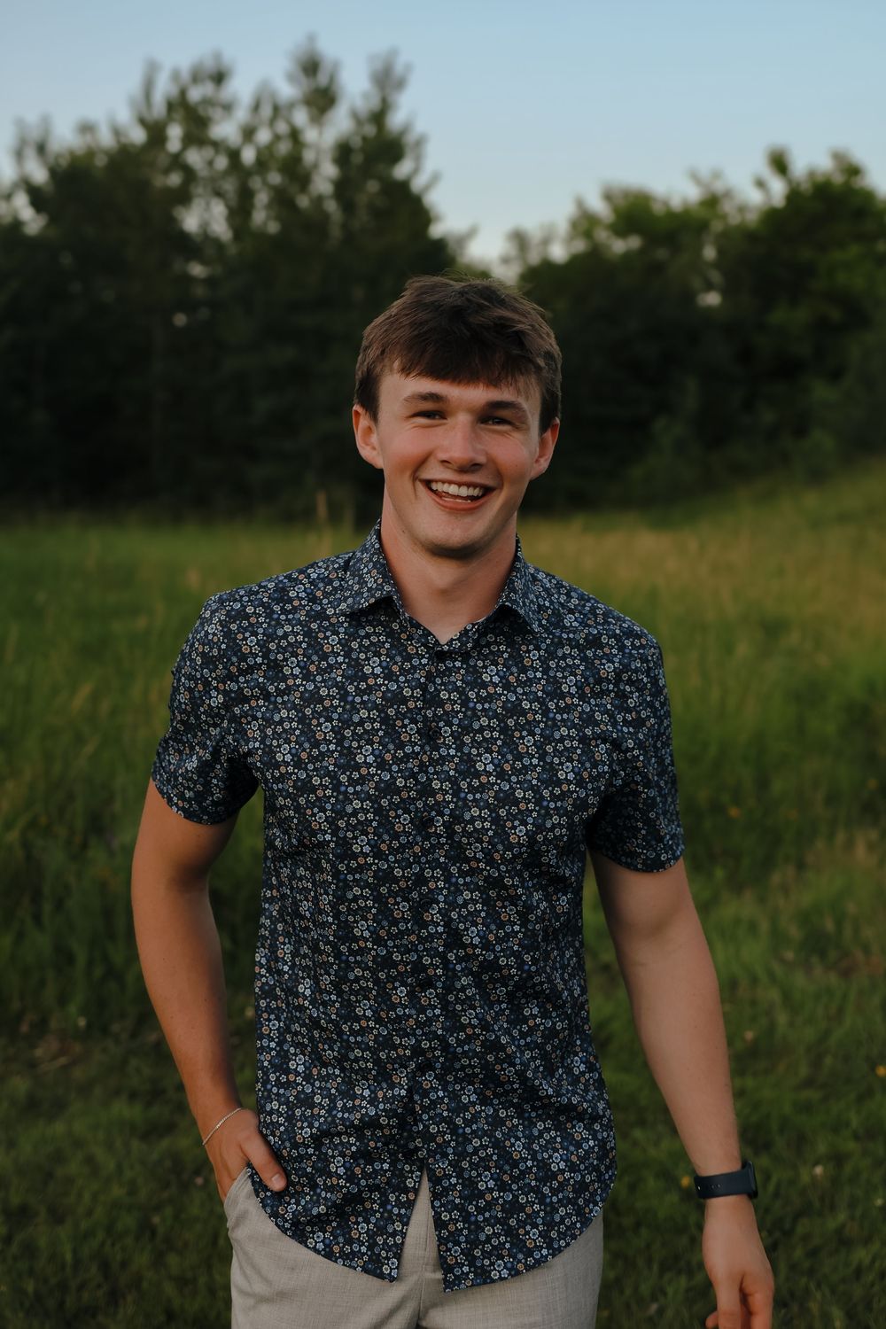 A candid outdoor portrait in a field with trees at sunset shows someone wearing a patterned short-sleeve button-down shirt.
