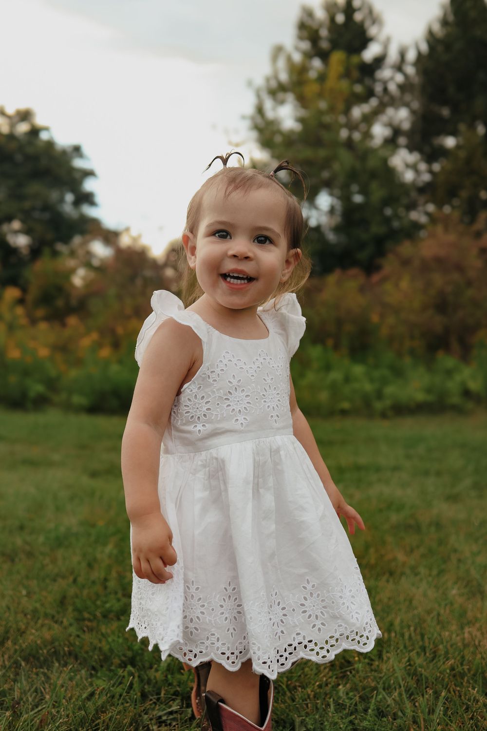 A small child in a white eyelet lace dress smiles while playing in a grassy field at sunset.